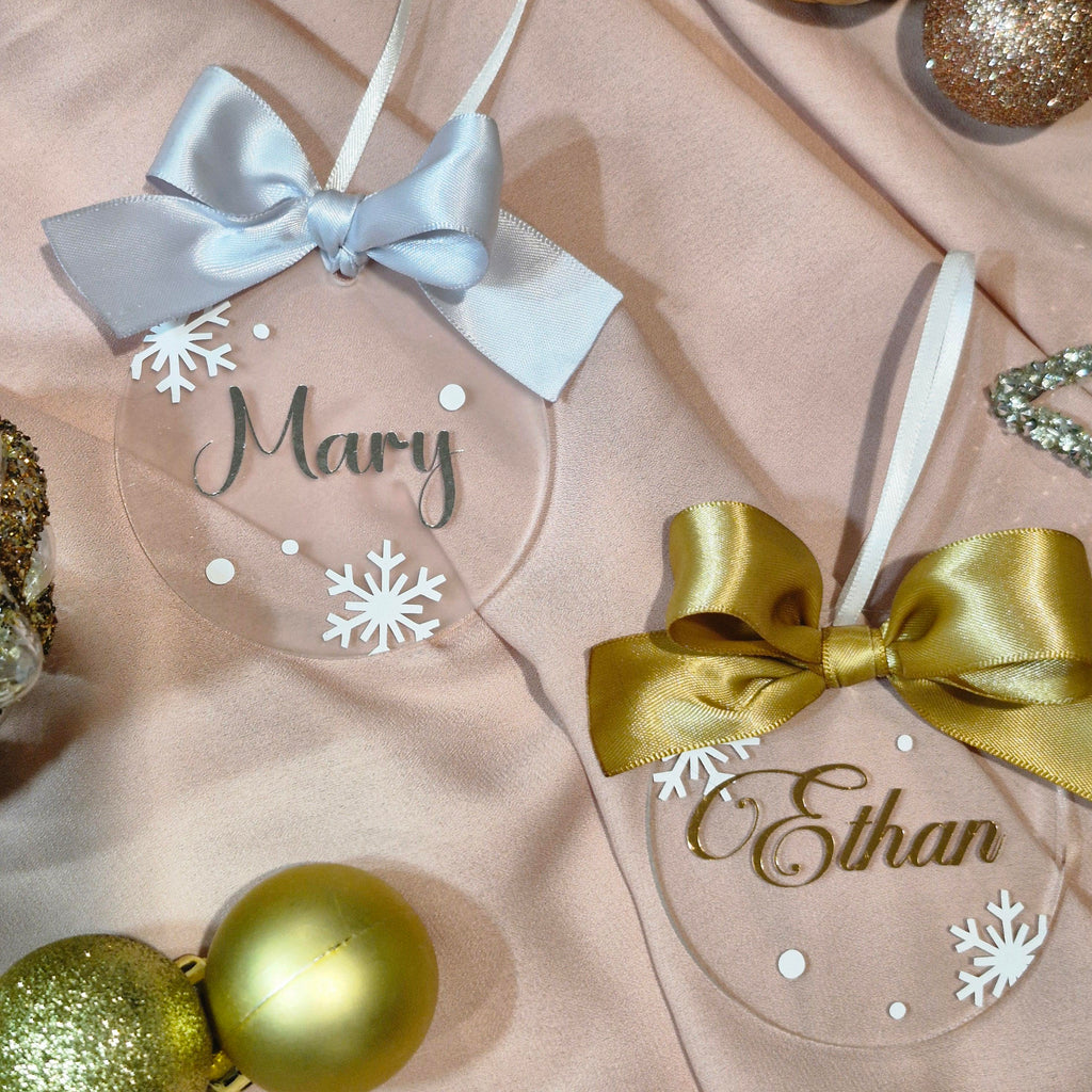 Two personalized clear acrylic Christmas ornaments with satin ribbon bows, displayed on a soft blush pink fabric background with gold and glitter ornaments. One ornament is labeled 'Mary' with a light blue bow, and the other is labeled 'Ethan' with a gold bow.