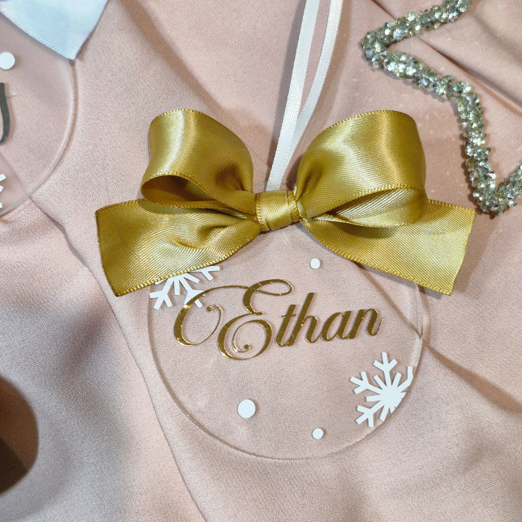 A close-up of a personalized Christmas ornament featuring a large gold satin bow, white snowflakes, and the name 'Ethan' in gold script on a pale pink fabric background.