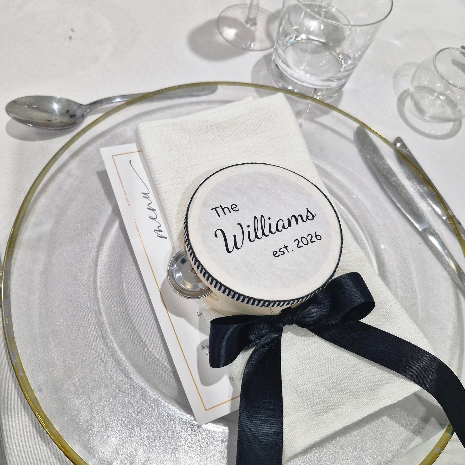 Personalized wedding favour tambourine with a black ribbon on a glass plate with glasses in the background.