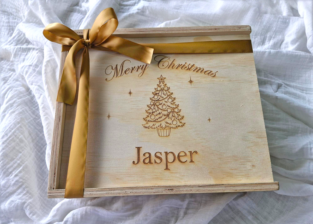 A personalized wooden Christmas gift box with a gold ribbon bow, featuring a laser-engraved Christmas tree and the name 'Jasper' on a white fabric background.