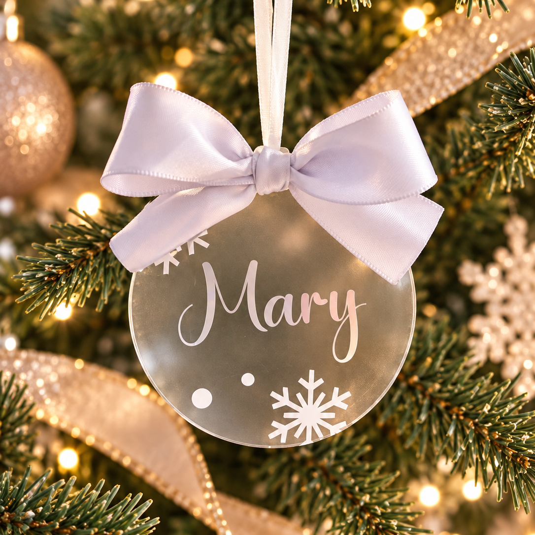 A round Christmas ornament with a white satin bow, hanging on a decorated tree. The ornament is frosted with the name 'Mary' in white script and white snowflake designs, surrounded by green pine branches and warm holiday lights.