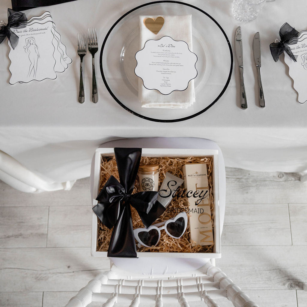 Wedding place setting with 'Bridesmaid' ribbon, glasses, and a box with sunglasses and bow.