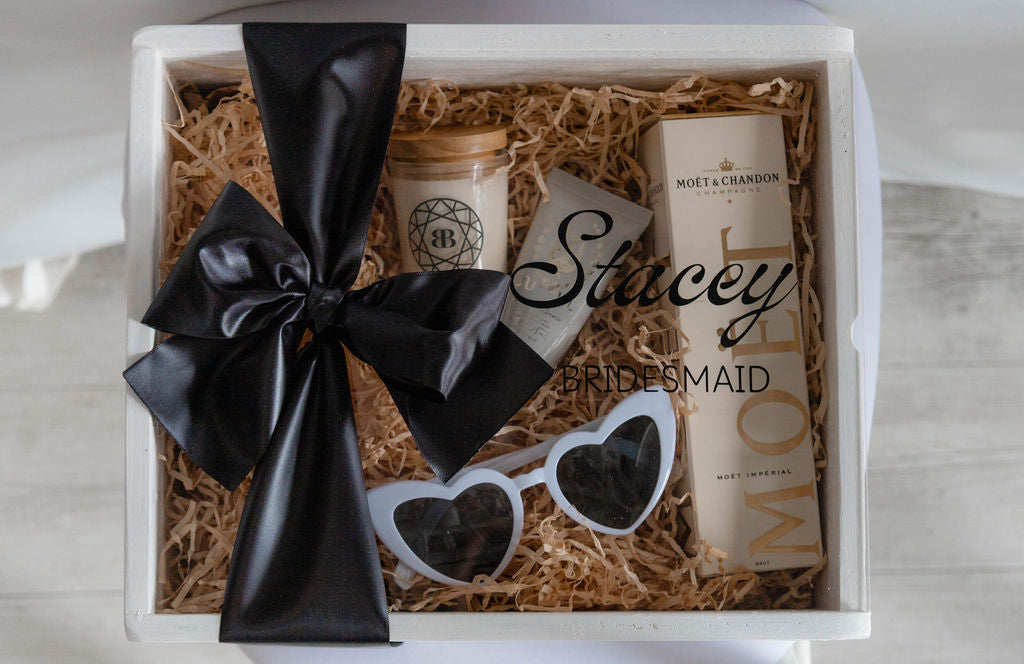 Gift box with a black bow, sunglasses, and a mini moet bottle, labeled 'Stacey Bridesmaid' on a light background.