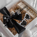 Gift box with black bow, heart-shaped sunglasses, and branded items on a white background