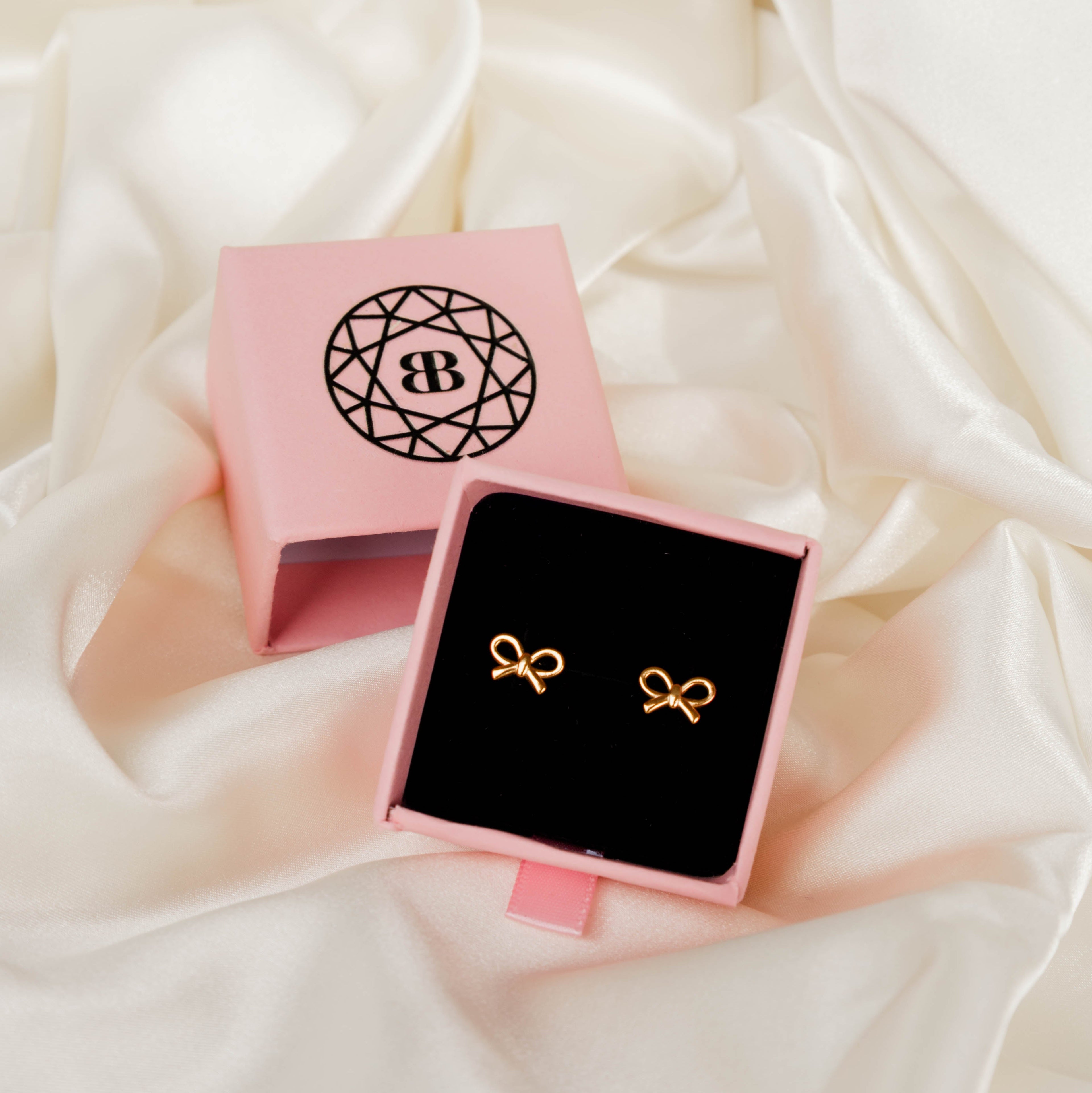 Gold bow-shaped earrings in a pink jewellery box on a white fabric background