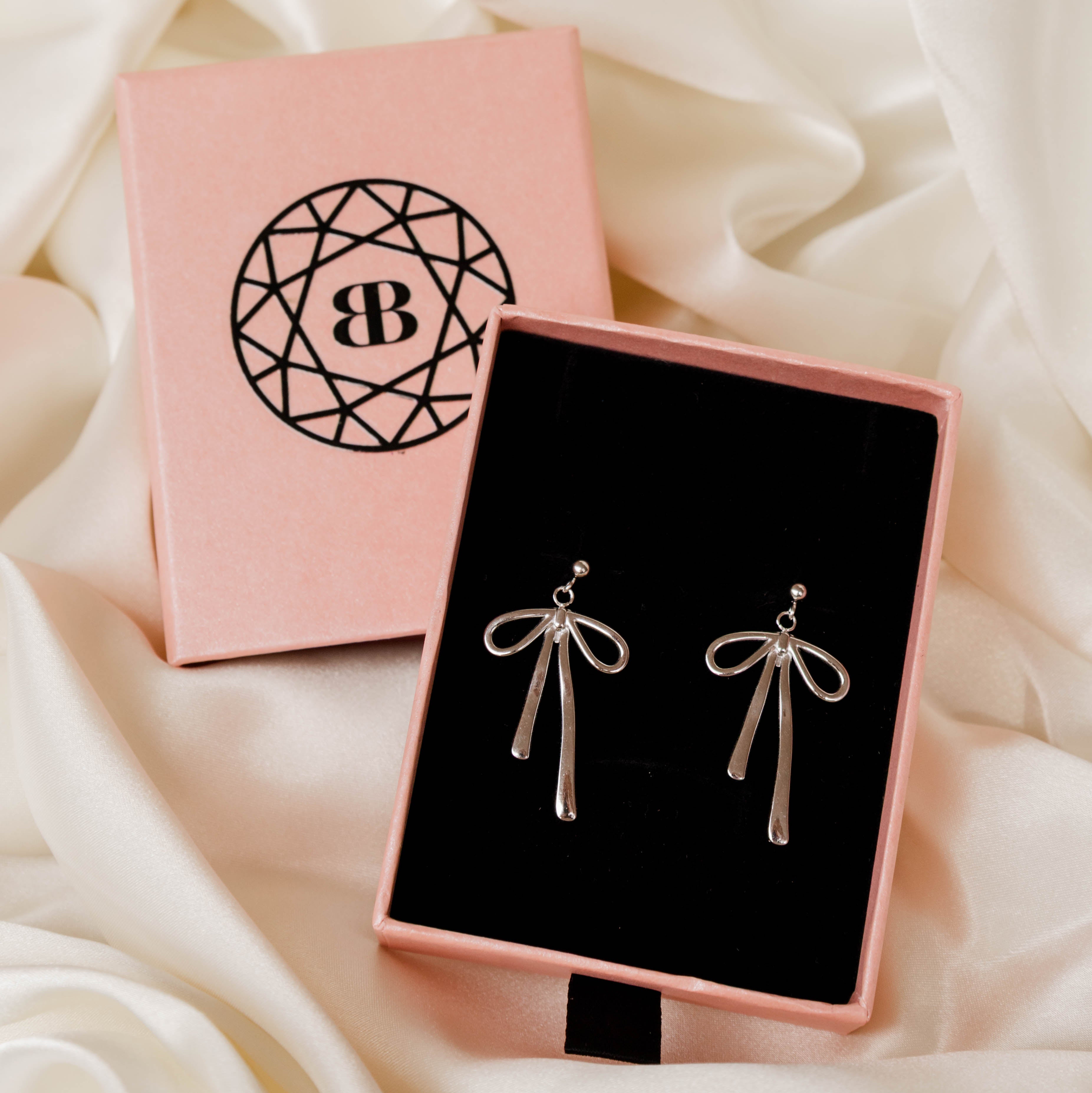 Silver bow-shaped earrings in a pink jewelry box with a brand logo on a soft white fabric background.