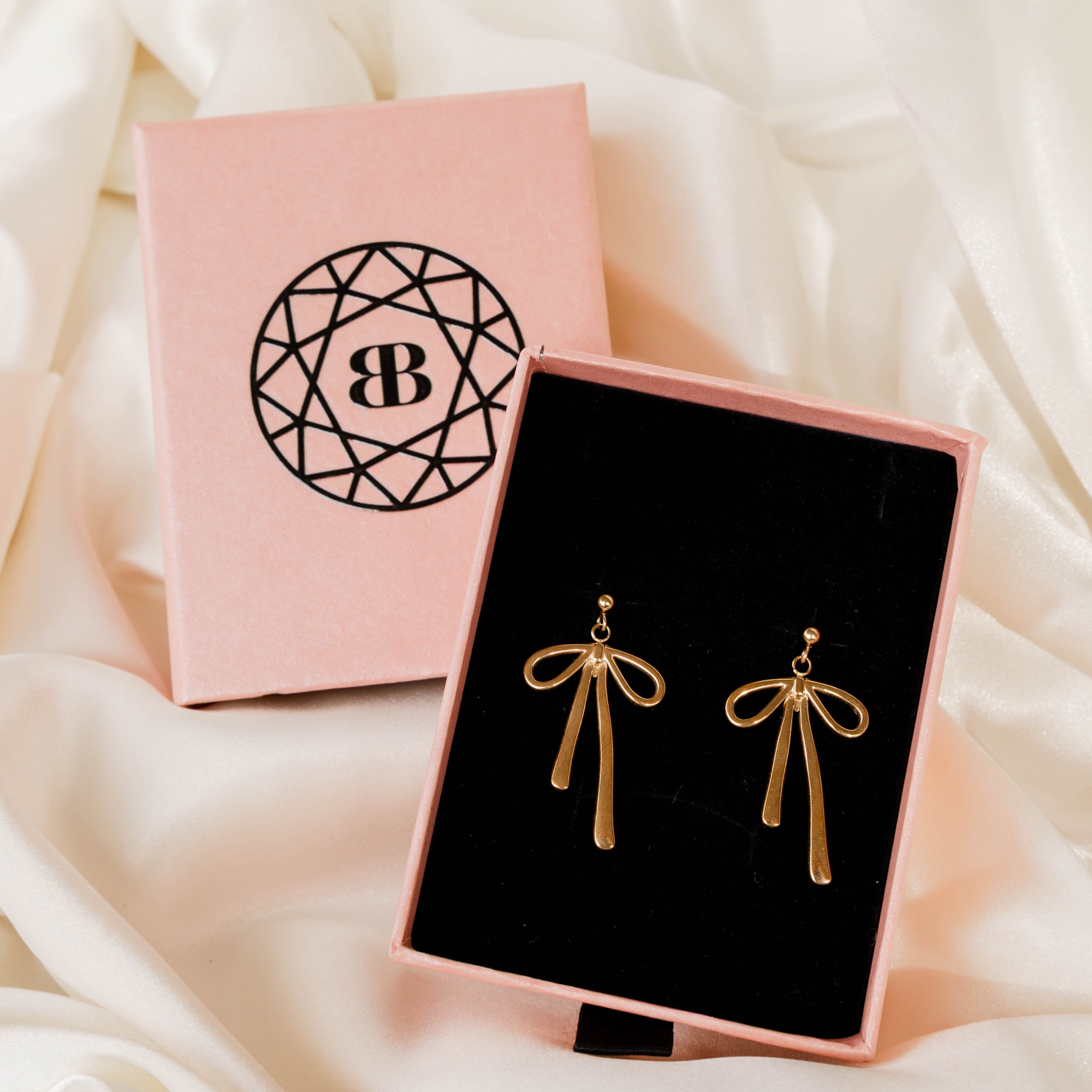 Gold bow-shaped earrings in a pink jewelry box with a visible brand logo on a soft white fabric background.