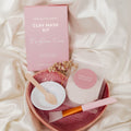 Clay mask kit with pink packaging, white mask, brush, and bowl on a soft fabric background
