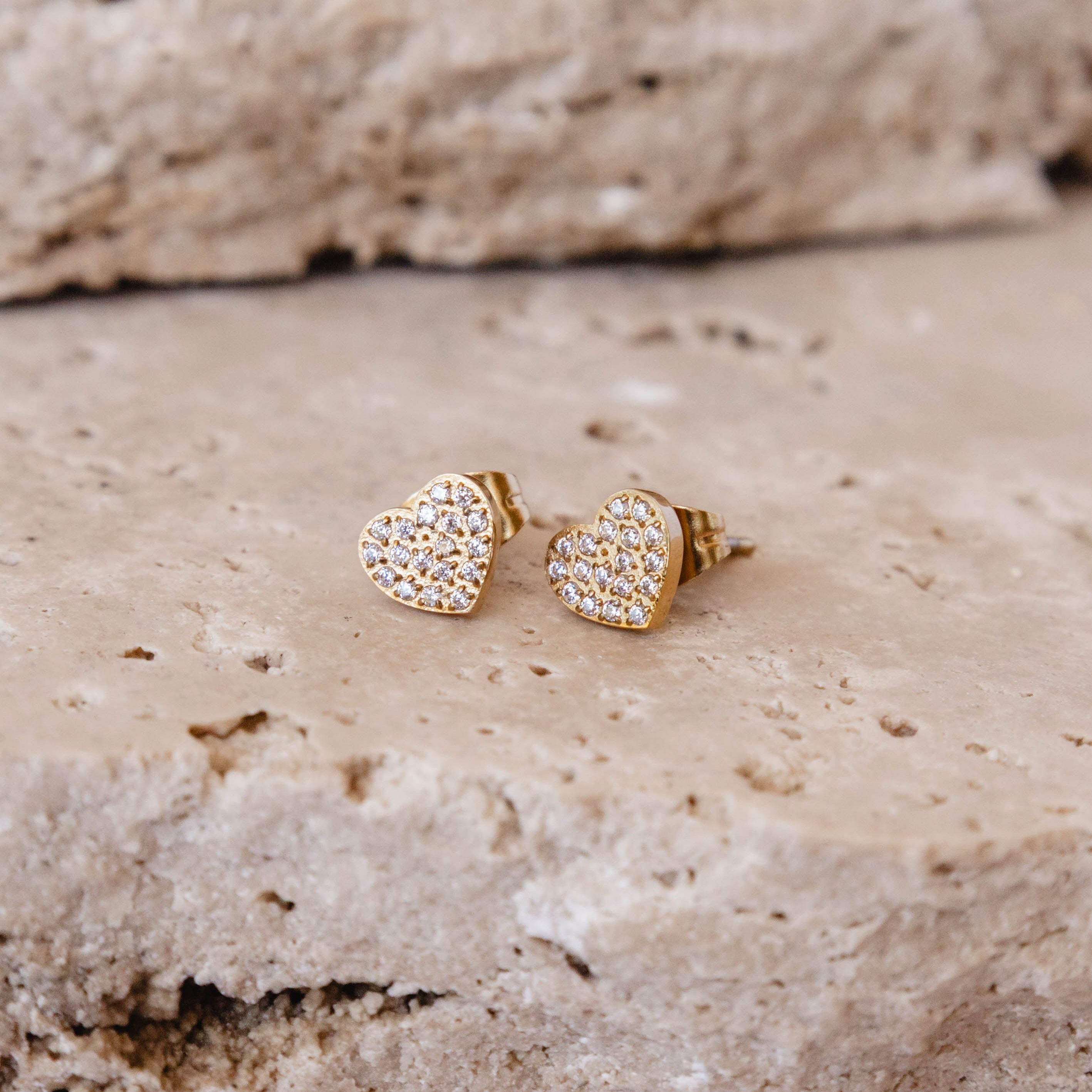 Heart-shaped gold earrings with clear stones on a textured stone surface