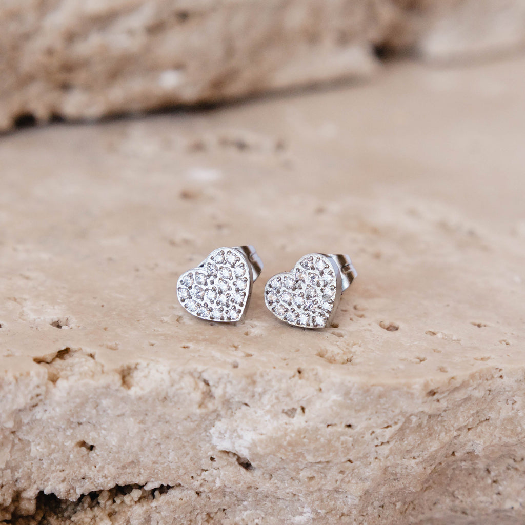 Heart-shaped silver earrings with clear stones on a beige stone surface