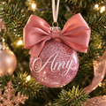 A round pink glitter Christmas ornament hanging on a decorated Christmas tree. The ornament features a large dusty pink satin bow on top and the name 'Amy' written in white cursive script on the front.