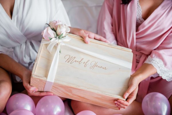 maid of honour wooden gift box with white ribbon and pink roses being handed between two women, one in a pink bridal robe and the other in a white robe