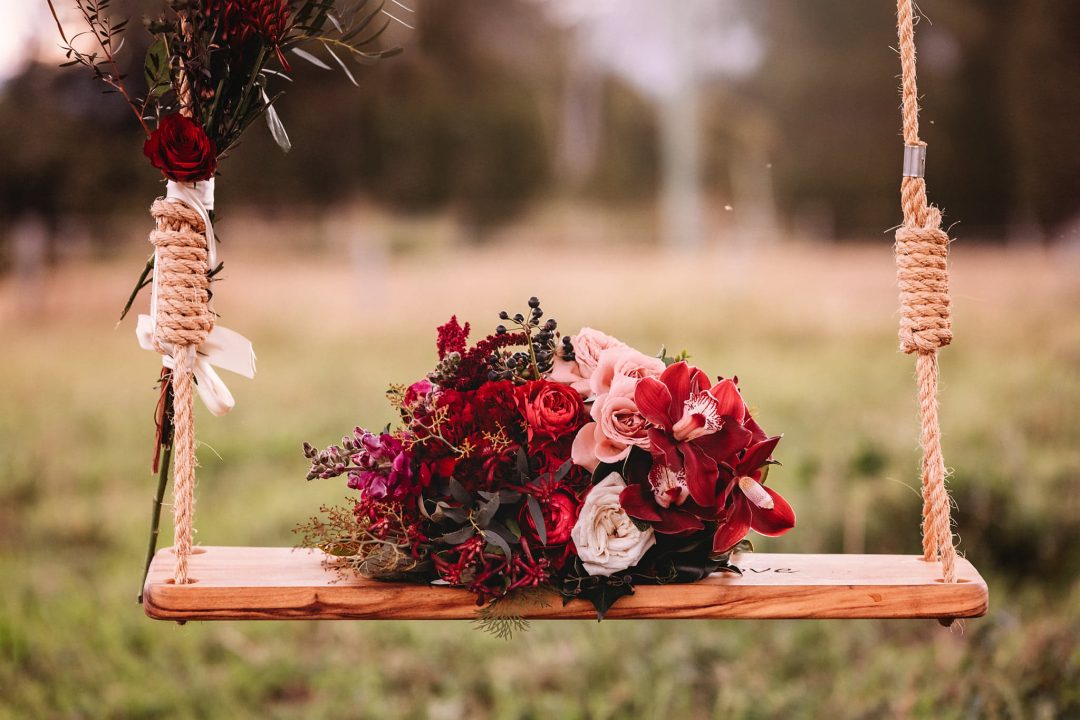 vibrant red and pink bouquet by Blooms by Bridie resting on a Bridal Box Co wooden swing