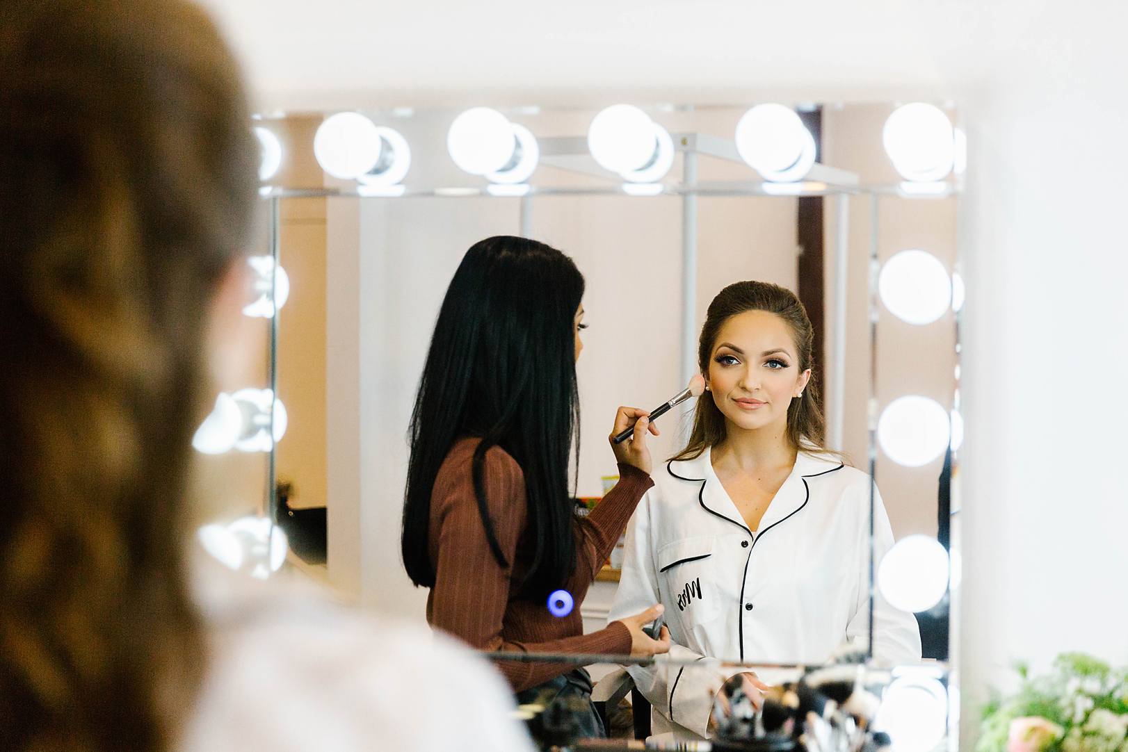 bride to be looking in illuminated mirror while getting her makeup done