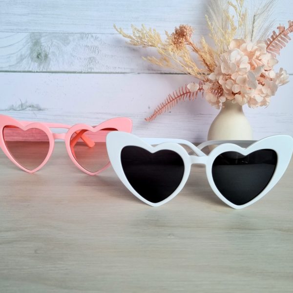a pair of retro heart sunglasses one in pink and one in white on tabletop with dried floral arrangement