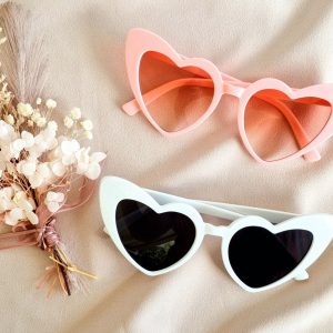 a pair of retro heart sunglasses one in pink and one in white on soft background with dried floral arrangement