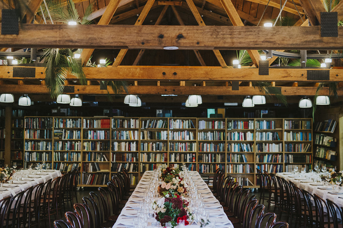 a barn like room, but with floral installations hanging from the ceiling and a large library bookshelt along the backwall
