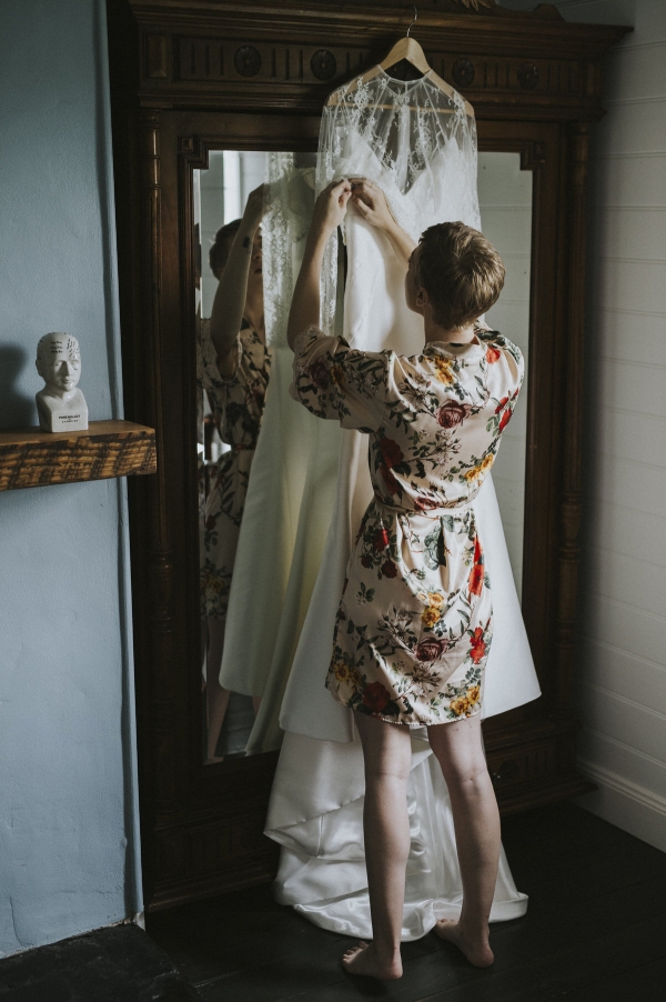 a bride standing in front of her wedding dress in a floral satin robe on the morning of her wedding