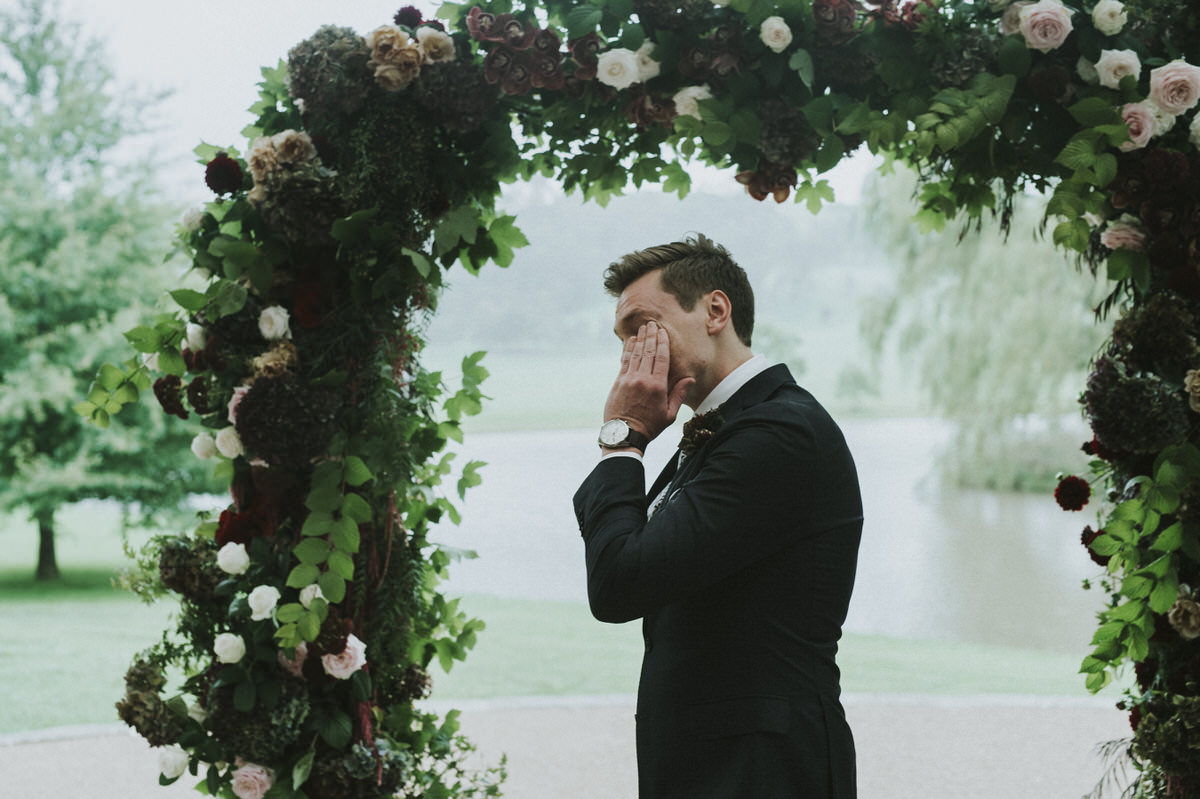 a groom crying and wiping tears from his eyes under a floral arbour