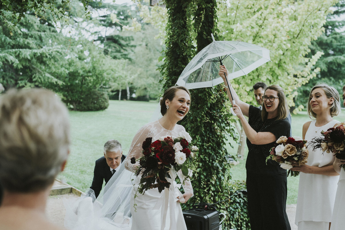 Bride laughing as she walks down the aisle in the rain and a bridesmaid is holding out an umbrella for her
