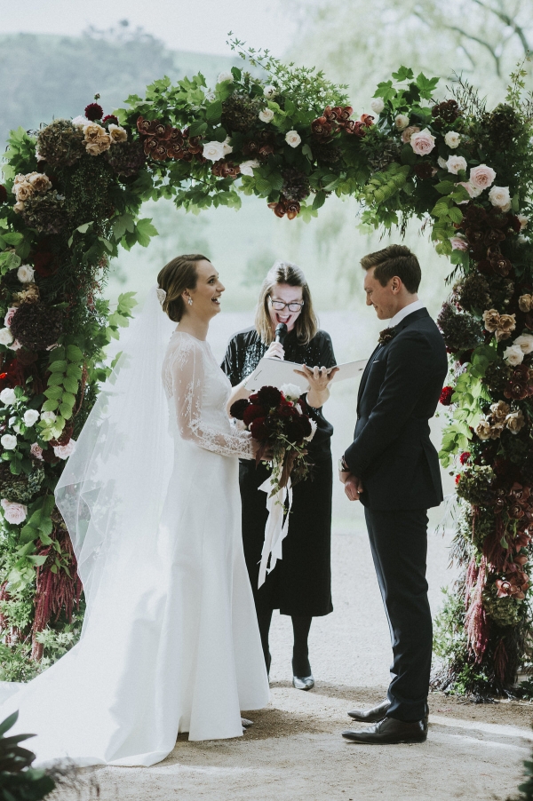 The bride and groom smiling and laughing as they exchange vows under a floral arbour