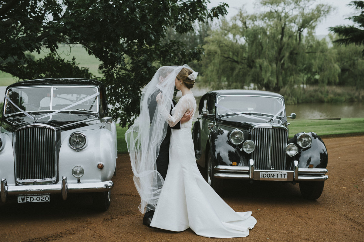 a bride and groom kissing in front of two old vintage cars, both under the brides veil