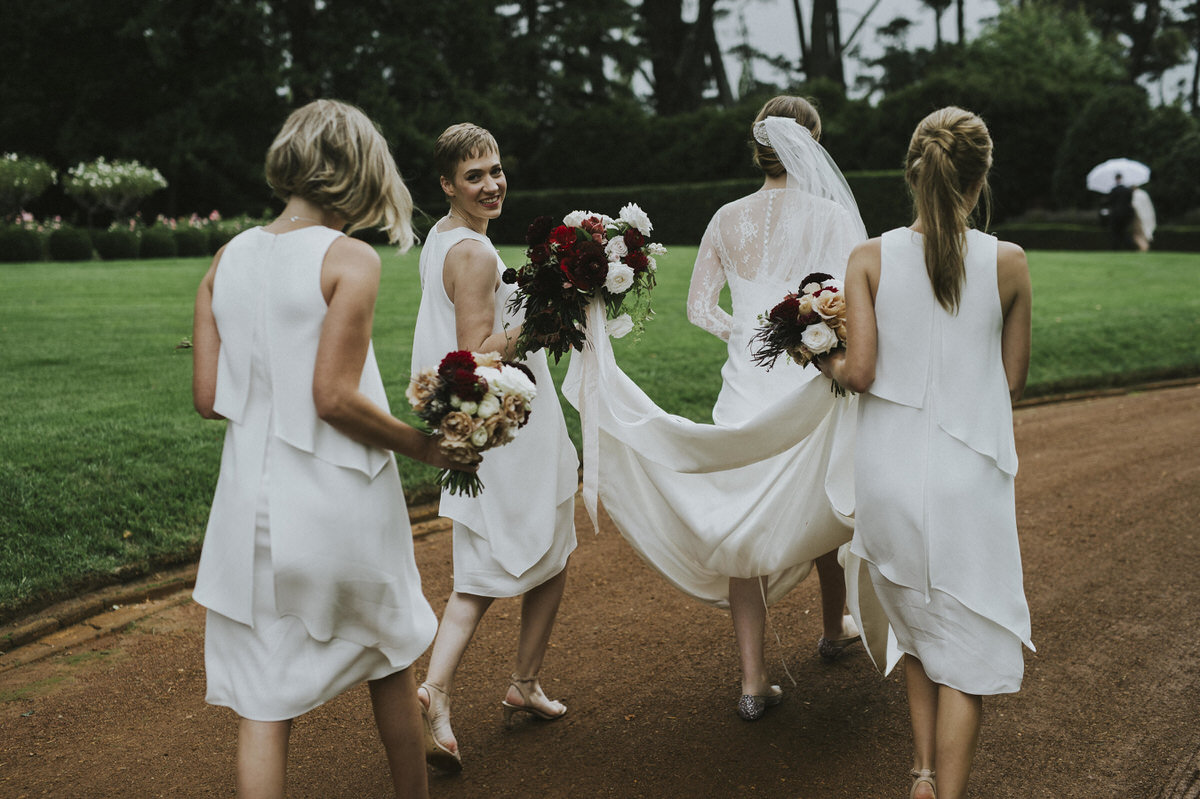 a bride and her bridesmaids walking down the road away from the camera. Everyone is dressed in white, walking along a red-dirt road. All but one is turned away from the camera