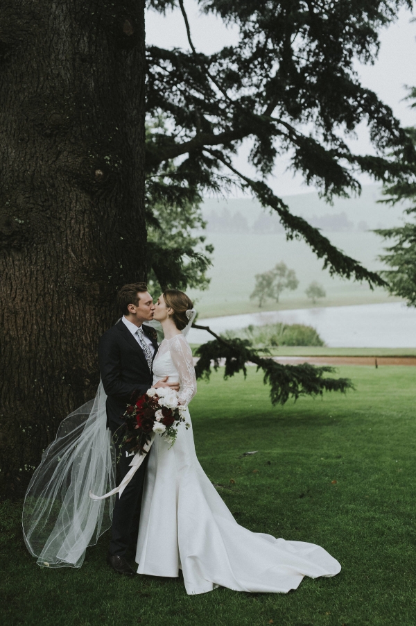 Bride and groom kissing under a large tree in the rain