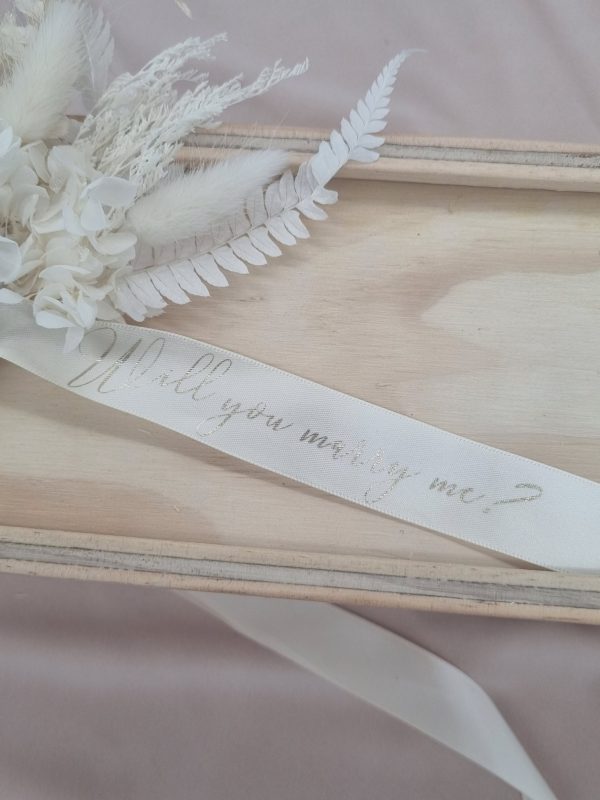 close up of Will You Marry Me? printed in gold foil on a white ribbon adorning a wooden keepsake box with dried floral arrangement